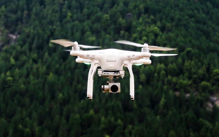 Weiße Kameradrohne im Flug über einem dichten grünen Wald.